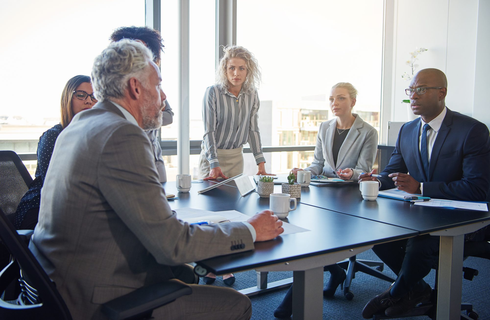 A board meeting by tall windows: a grey-haired director speaks while the others at the table listen