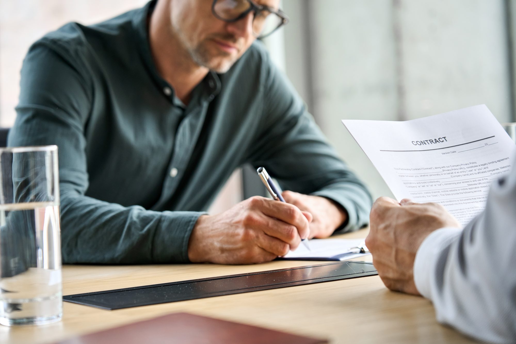 A man in glasses signing a contract while the other party holds the document